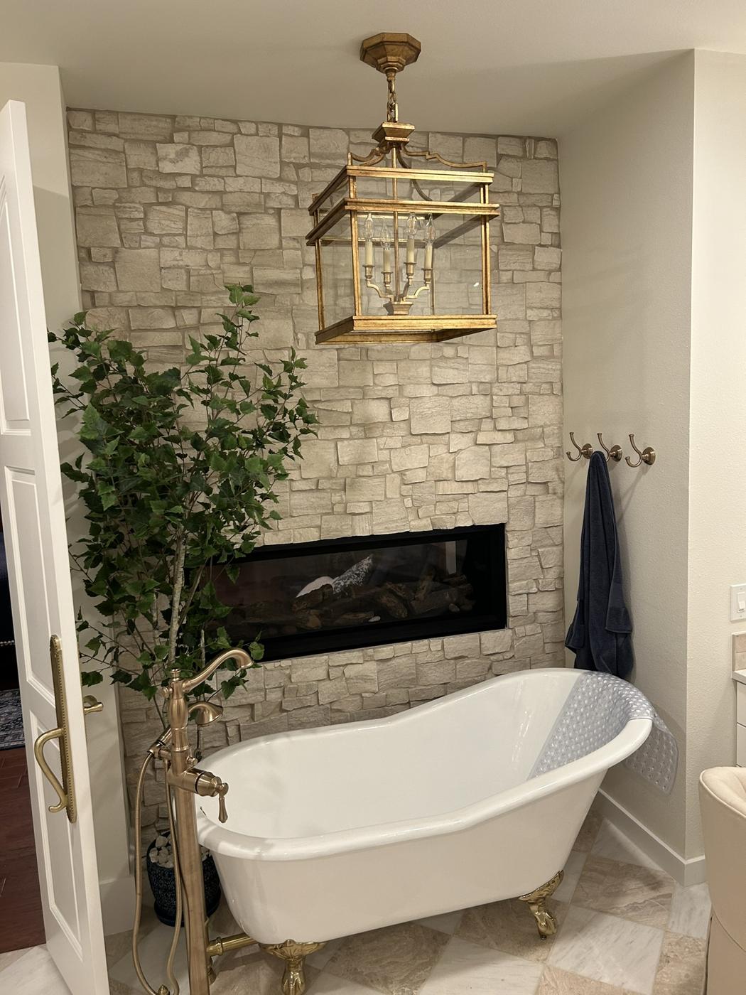 Freestanding clawfoot tub in front of a stacked-stone wall with a linear gas fireplace, brass chandelier above